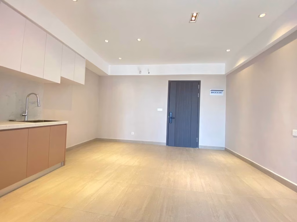 A bright, empty room at Time Square 3 Condo features beige tile flooring, white walls, a dark wooden door, recessed ceiling lights, and a small kitchen area with a sink and cabinets on the left.