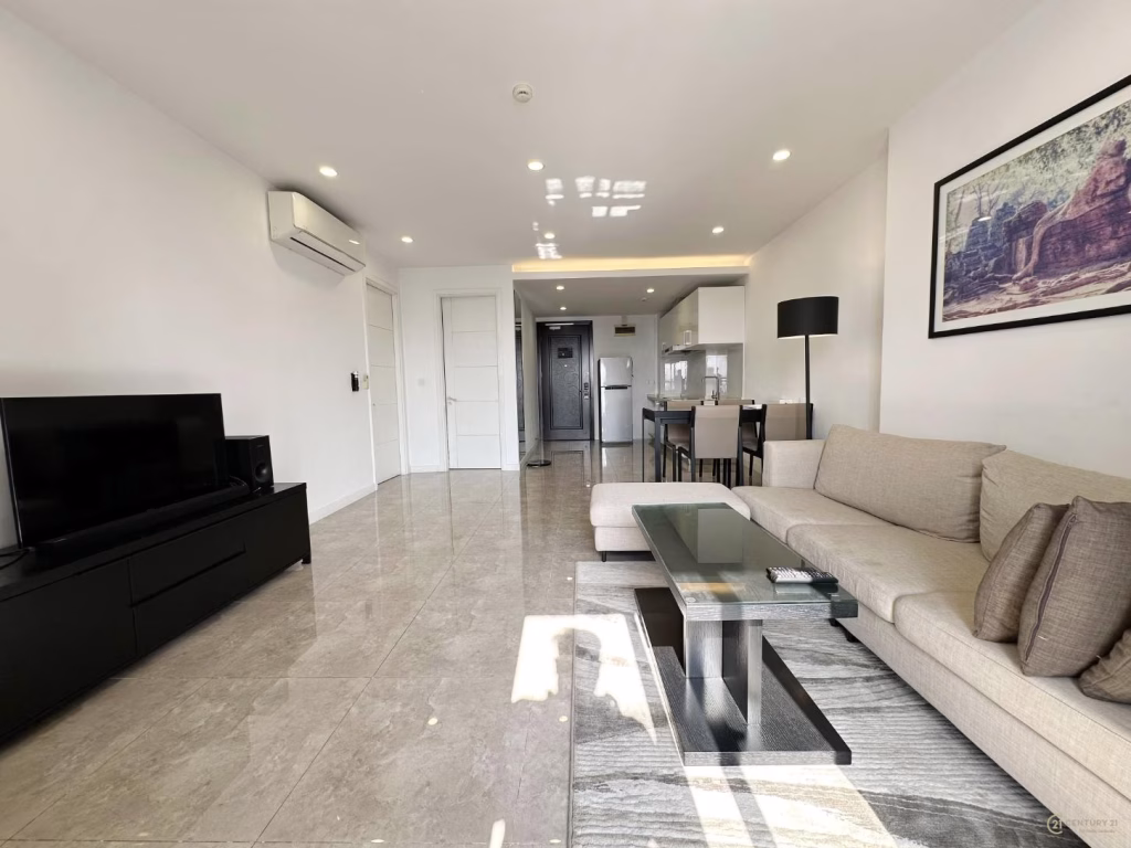 Modern condo 2 bedroom living room with a beige sectional sofa, glass coffee table, floor lamp, flat-screen TV on a black stand, and an open kitchen in the background. Large tile flooring and minimalistic decor.