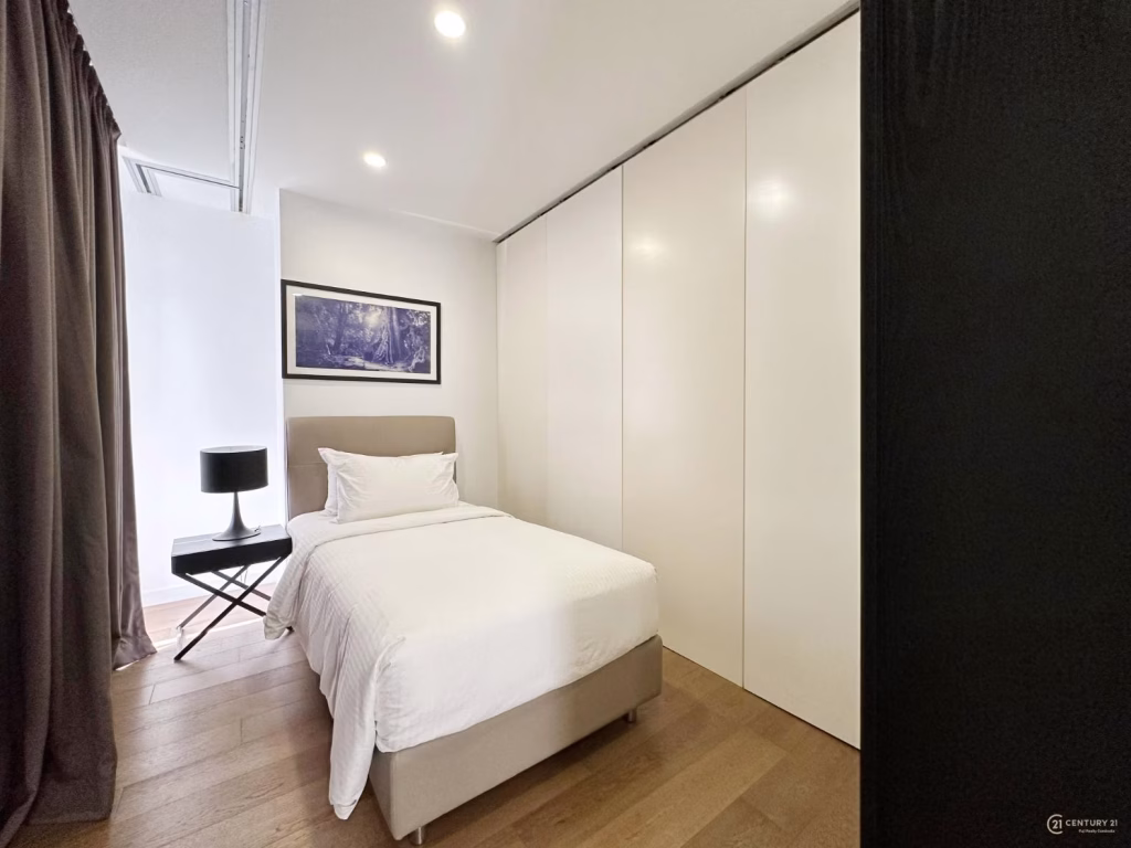 A modern, minimalist condo 2 bedroom features a single bed with white linens, a side table with a black lamp, floor-to-ceiling closets, wood flooring, and a framed landscape photo on the wall.