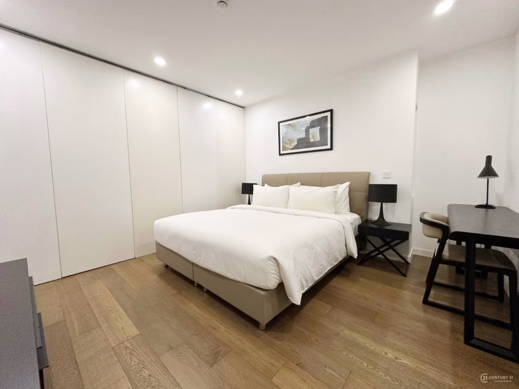 Modern Condo 2 Bedroom with a large bed, white bedding, two black lamps on bedside tables, a desk with chair and lamp, wooden floor, and white built-in wardrobes. A framed map hangs above the bed.