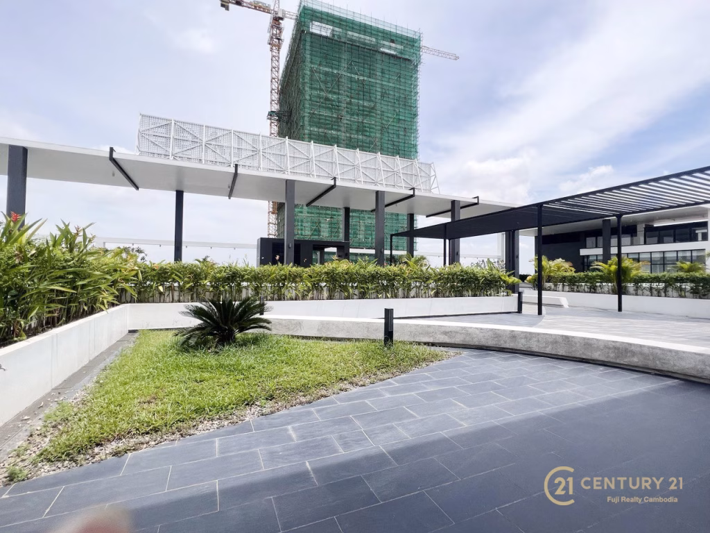 A modern outdoor area with landscaped greenery, tiled walkways, and shaded structures. In the background, Urban Village Phnom Penh rises under green scaffolding. The Century 21 logo is visible in the bottom right corner.
