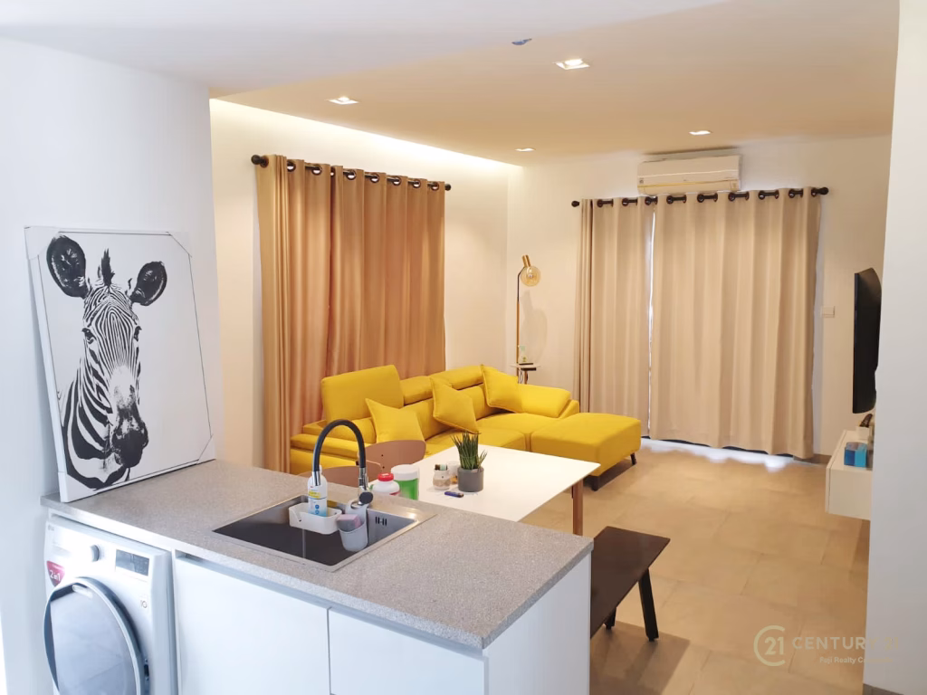 Modern living room at Urban Village Phnom Penh featuring a yellow sectional sofa, beige curtains, white walls, and a zebra portrait above a washing machine and kitchen sink. Natural light fills the contemporary space.