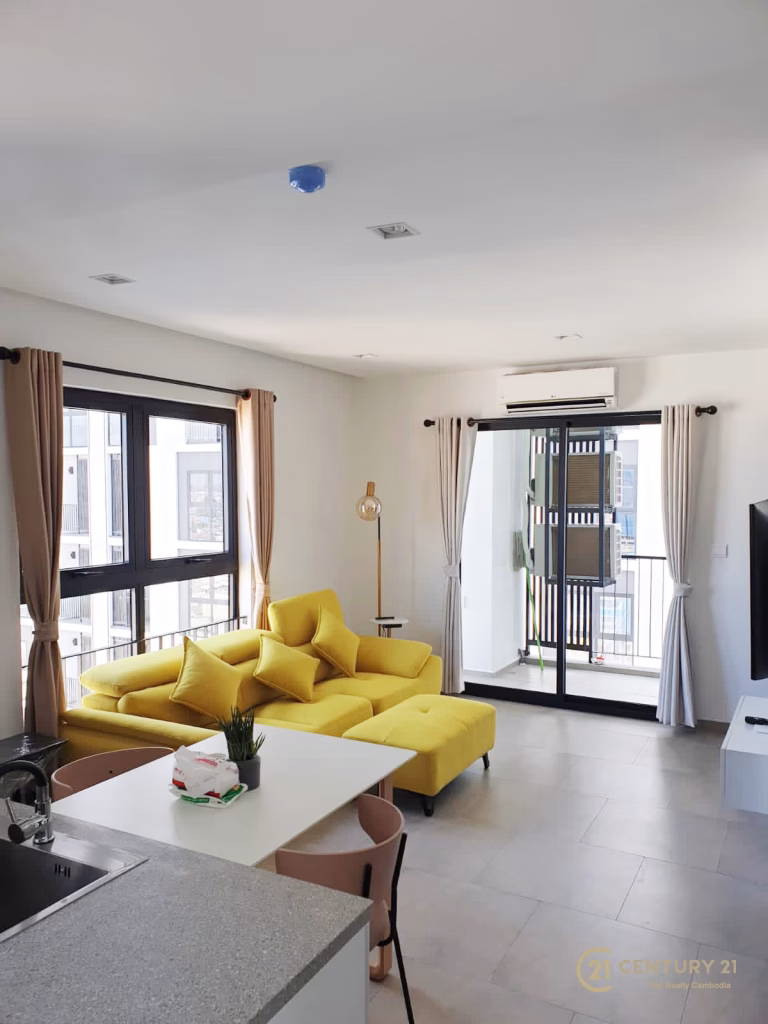 A modern living room in Urban Village Phnom Penh with large windows, beige curtains, a bright yellow sectional sofa, a small dining table, and a sliding glass door leading to a balcony. The space is bright and neatly arranged.