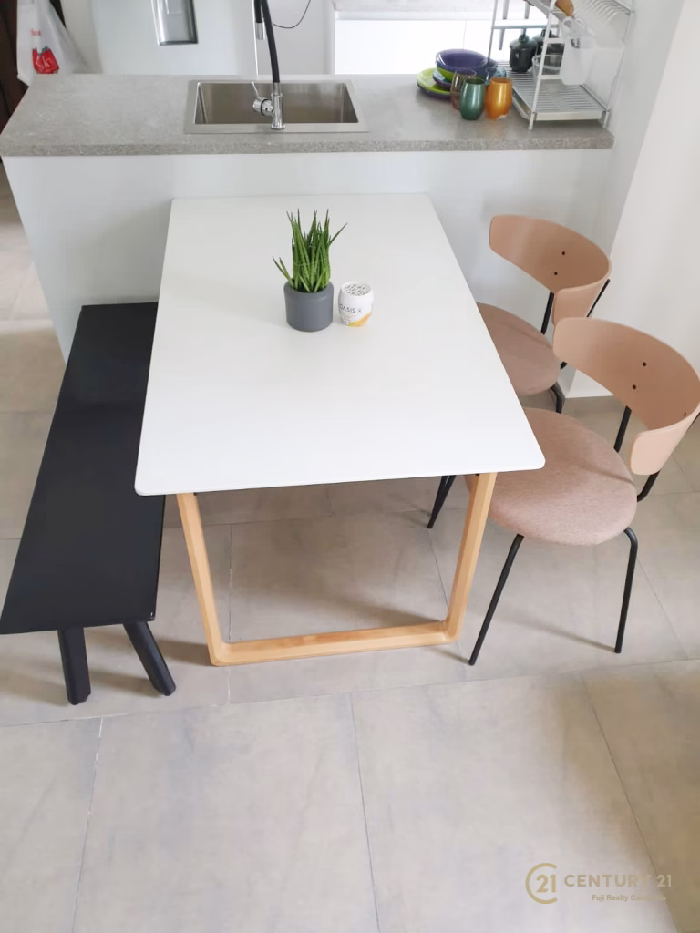 A modern dining area at Urban Village Phnom Penh features a white rectangular table, a black bench, beige chairs, and décor like a potted plant and candle. The kitchen sink and dish rack are visible in the background.