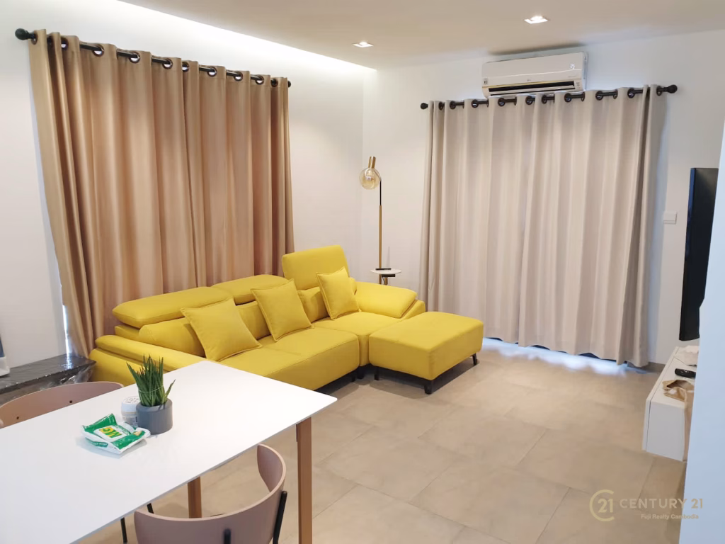 Modern living room at Urban Village Phnom Penh featuring a bright yellow sectional sofa, beige curtains, a small round side table with a lamp, and a wall-mounted air conditioner. A dining table with chairs sits in the foreground.