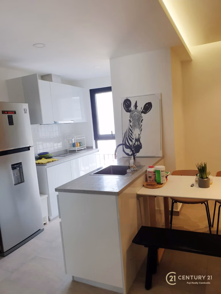 A modern kitchen with white cabinets, a stainless steel refrigerator, a small dining table with chairs and a bench, and zebra artwork on the wall near the window—perfect for Urban Village Phnom Penh living. Image branded with the Century 21 logo.