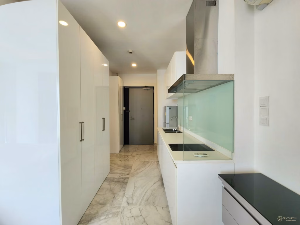 Modern, narrow kitchen in a condo for sale with white cabinets, marble floor, glossy surfaces, built-in appliances, glass backsplash, electric cooktop, and stainless steel vent hood under bright ceiling lights. A gray door is visible at the end.