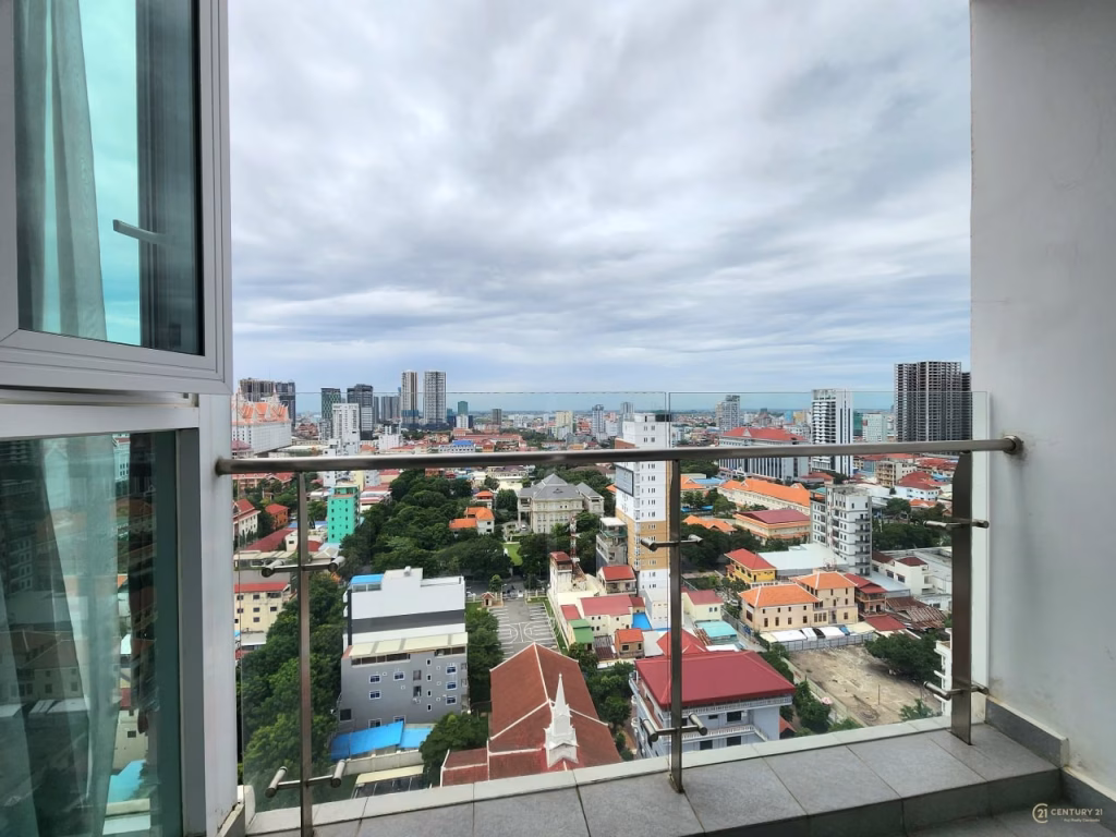 A cityscape view from a high balcony of a condo for sale, featuring modern buildings, colorful rooftops, and the ocean in the distance under a cloudy sky. Glass railing lines the balcony.