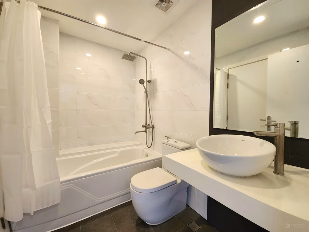 Modern bathroom in this condo for sale features white tiled walls, a bathtub with rain showerhead, a vessel sink on a countertop, and a large mirror. The bright lighting and white shower curtain enhance the fresh, contemporary feel.