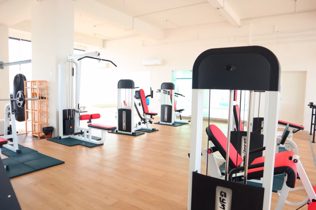 A bright, spacious gym in this condo for sale features wooden floors and large windows, with various exercise machines boasting black, white, and red padding, including weight machines and benches.