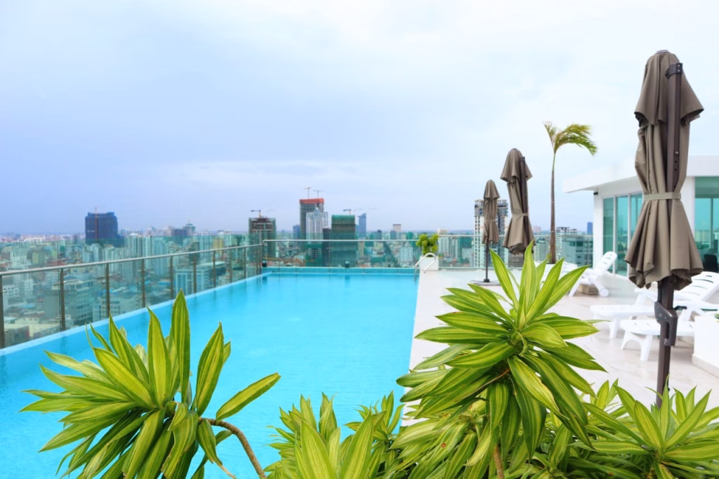 A rooftop swimming pool with clear blue water, surrounded by green plants and closed umbrellas, overlooks a city skyline under a bright, cloudy sky. White lounge chairs line the deck—luxurious amenities for this stunning condo for sale.