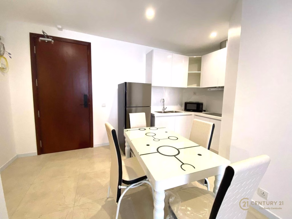Modern kitchen and dining area at The Penthouse Residence with white walls, tiled floor, a rectangular patterned glass dining table and four chairs, stainless steel fridge, microwave, white cabinets, and a wooden door in the background.
