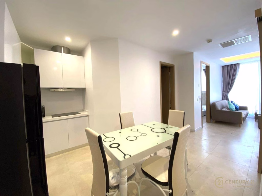 Modern apartment interior at The Penthouse Residence, featuring a small white kitchen, glass dining table with four chairs, and a living area with a sofa near a large window with curtains. Bright, minimalist design.