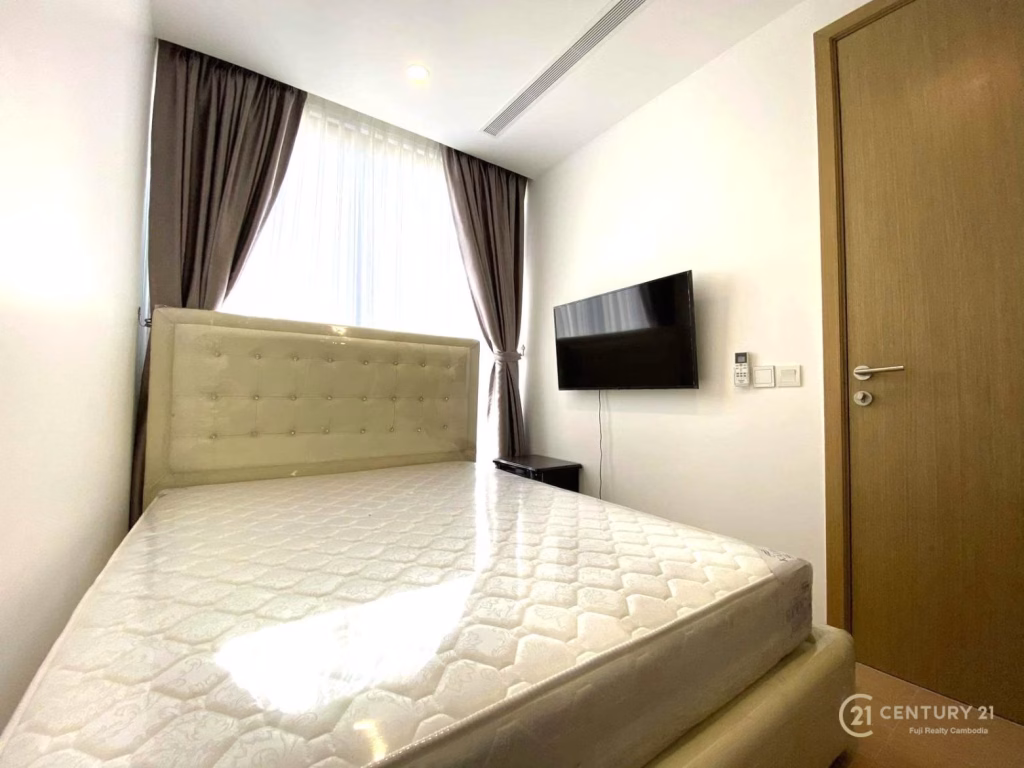A small, modern bedroom at The Penthouse Residence features a bed frame and mattress, floor-to-ceiling window with sheer and dark curtains, wall-mounted TV, wooden door, and white walls. The room is bright and minimalistic.