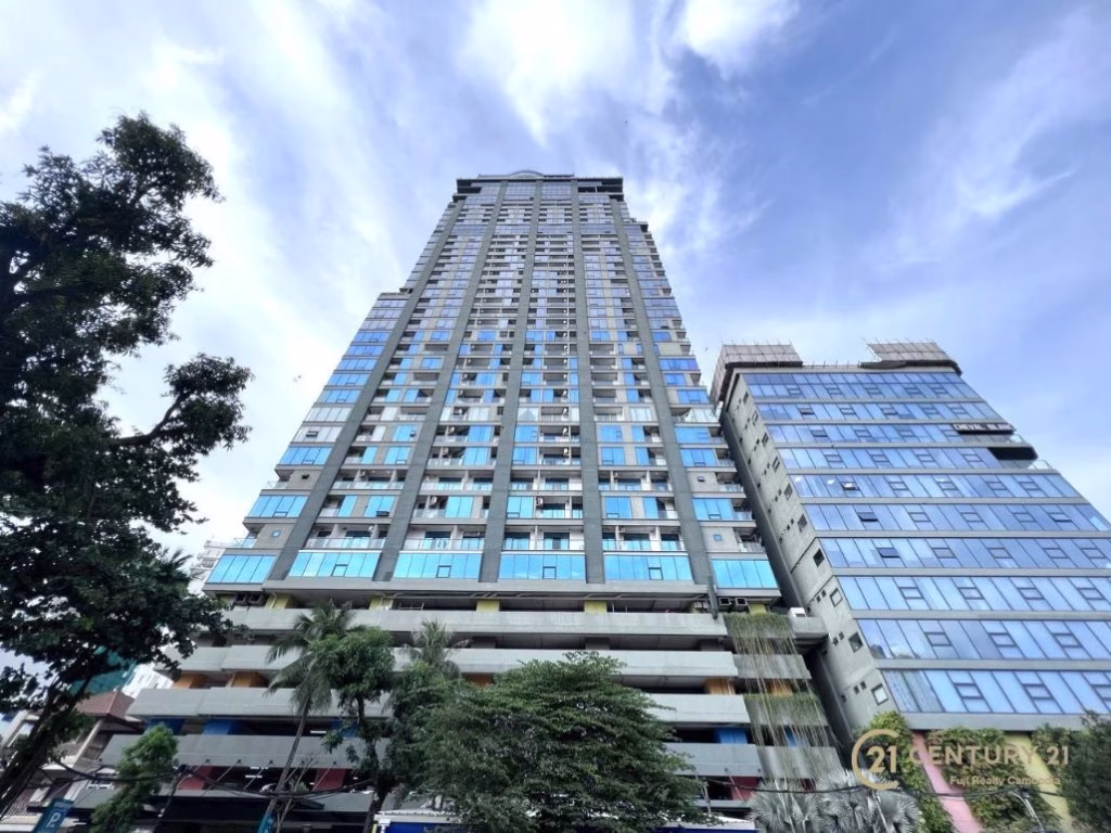 A tall modern high-rise, home to The Penthouse Residence, features blue-tinted windows and rises against a partly cloudy sky, with trees and another building in the foreground.