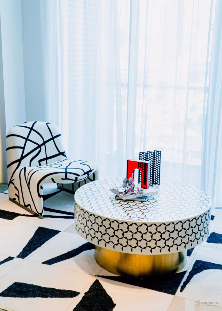 A modern living room from the Time Square 6 Project features a geometric-patterned chair, a round coffee table with a white and black design on a gold base, and decorative items; sheer curtains filter natural light in the background.
