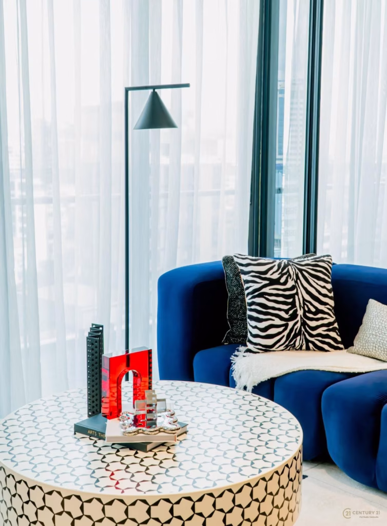 A modern living room corner styled for the Time Square 6 Project features a blue armchair, zebra-print pillow, and white throw blanket. A round patterned coffee table with decor sits beside a tall black floor lamp and sheer curtains drape tall windows.