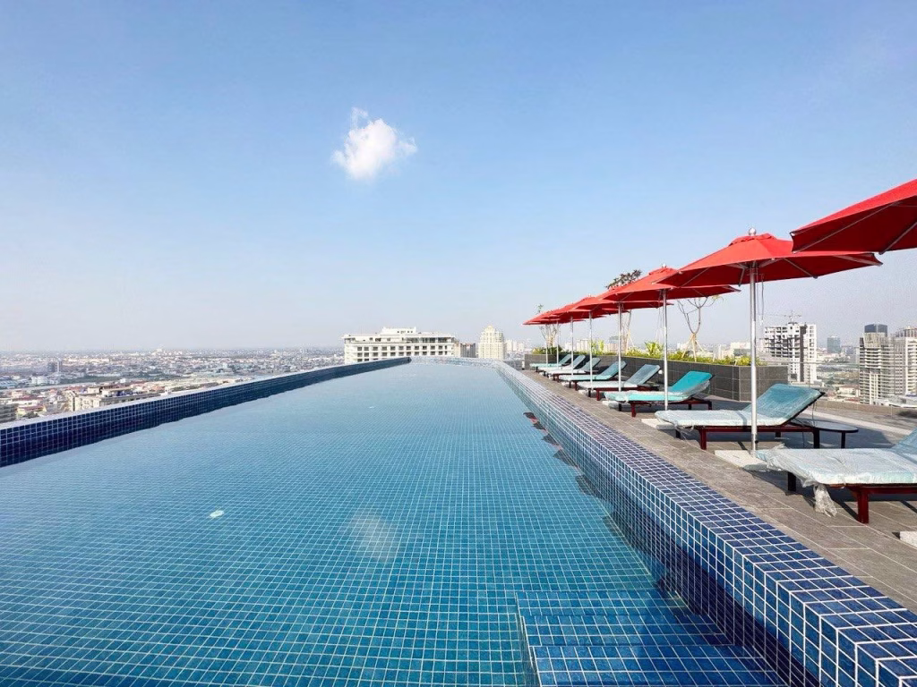 A rooftop infinity pool at Time Square 3 overlooks a cityscape under a clear blue sky. Red umbrellas shade lounge chairs along the poolside, creating a relaxing atmosphere for sunbathing and leisure.
