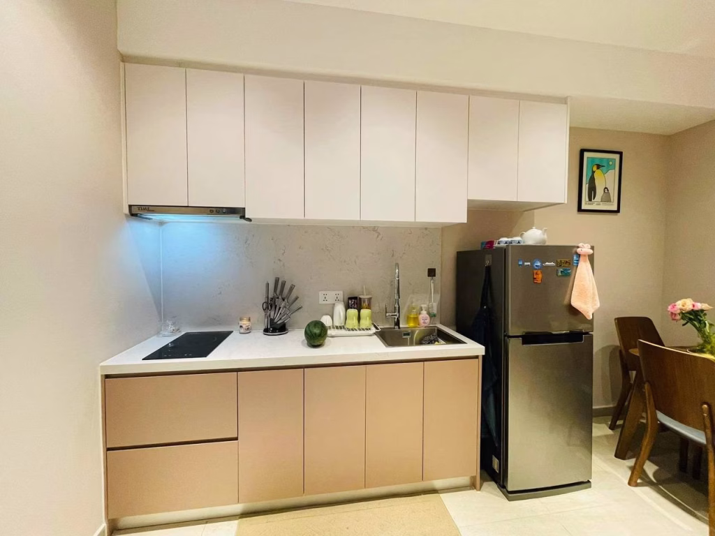 A modern kitchen at Time Square 3 with beige lower cabinets, white uppers, a sink, stove, knife set, and utensils on the counter beside a silver refrigerator. A small dining table with flowers and a framed picture are nearby.