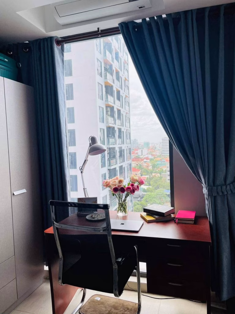A modern home office setup with a dark wooden desk, black chair, laptop, lamp, books, and a vase of flowers beside a large window with blue curtains offers a cityscape view inspired by Time Square 3.