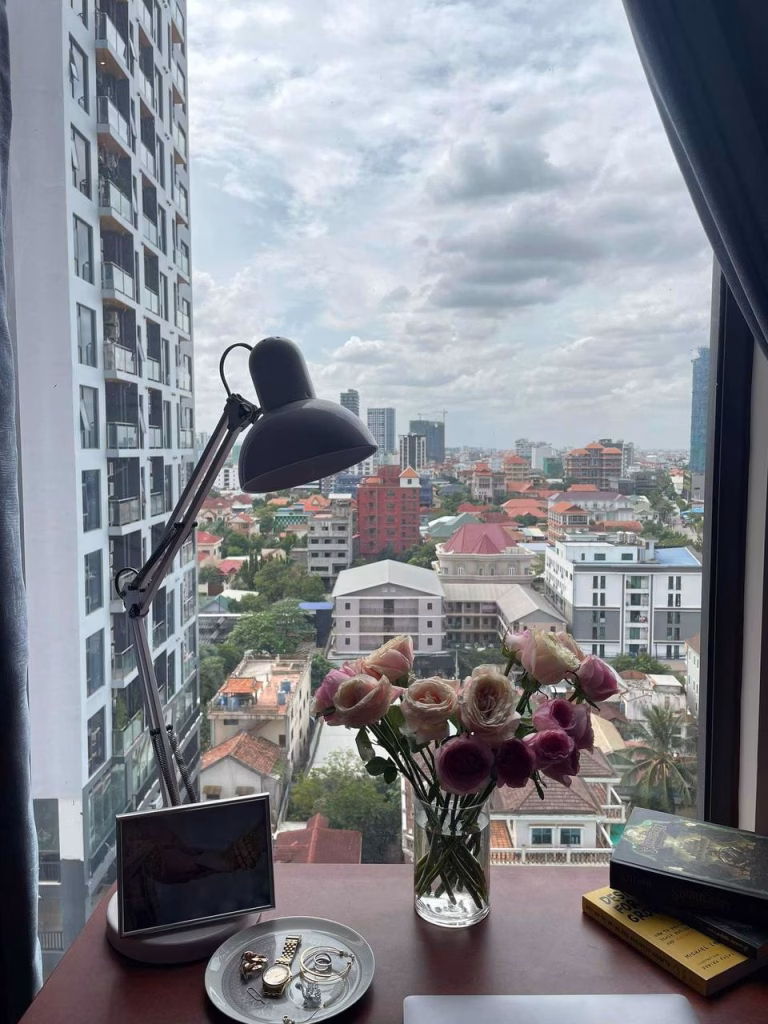 A desk by a window with a city view, featuring a black lamp, a vase of pink and white flowers, a photo frame, jewelry on a plate, and books—perfectly capturing the vibe of Time Square 3 under a cloudy sky.
