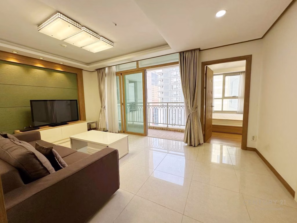 Modern living room at De Castle Royal BKK1 with a brown sofa, TV on a white stand, coffee table, large windows with curtains, and a balcony. An adjacent bedroom is visible through an open sliding door. The space is bright and tidy.