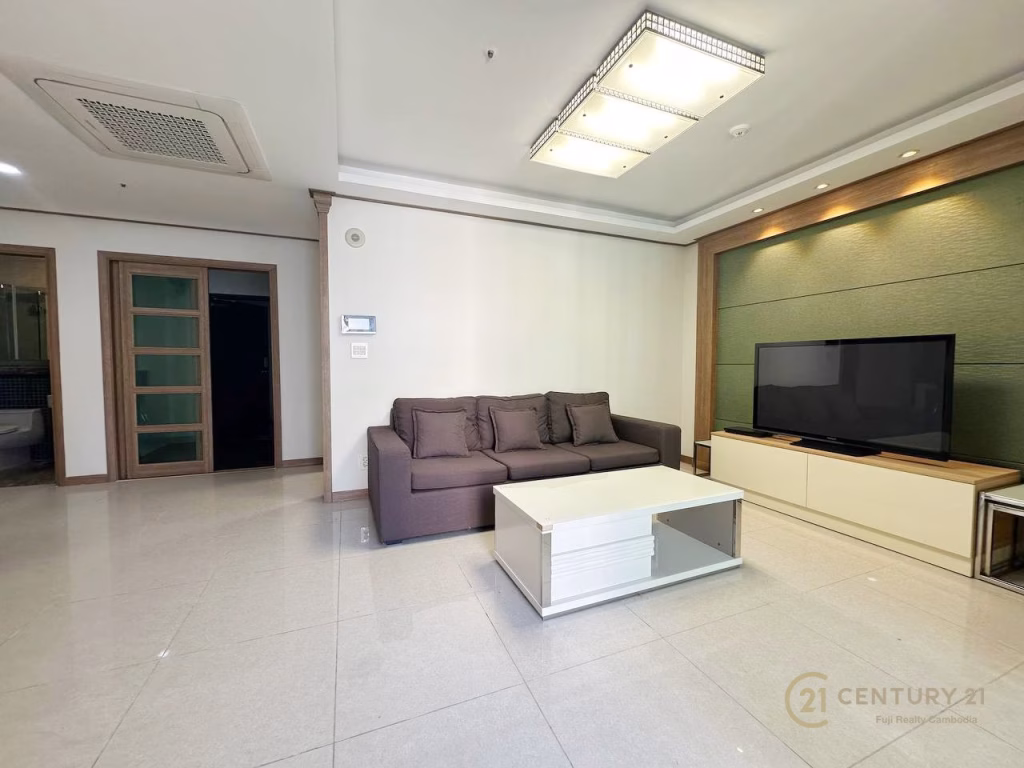 Modern living room at De Castle Royal BKK1 featuring a gray sofa, white coffee table, and flat-screen TV on a light-colored stand. The space is bright with tiled floors, recessed lighting, and doors leading to a bathroom in the background.