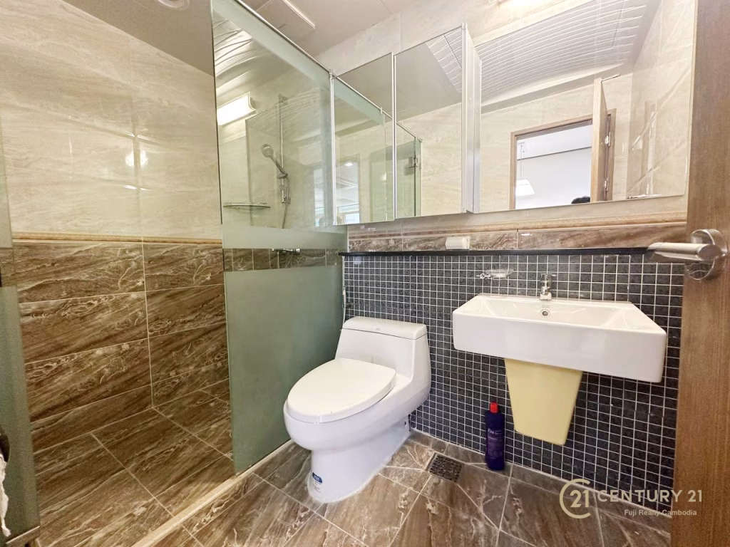 Modern bathroom at De Castle Royal BKK1 with marble-effect tiles, a glass shower enclosure, a white toilet, a wall-mounted sink, mirror, and toiletries on the floor; Century 21 logo in the corner.