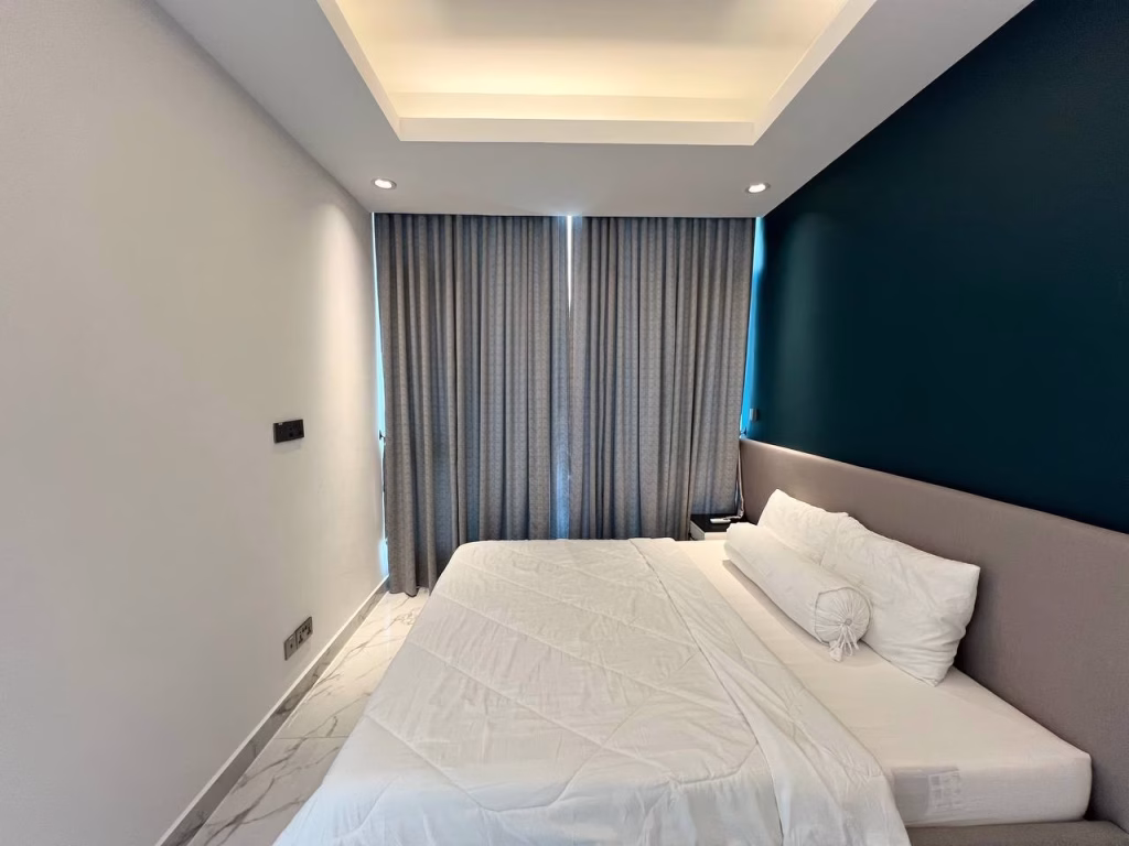 Modern 2 bedroom condo featuring a double bed with white sheets, gray curtains over the window, a teal accent wall, ceiling lights, and elegant marble tile flooring.