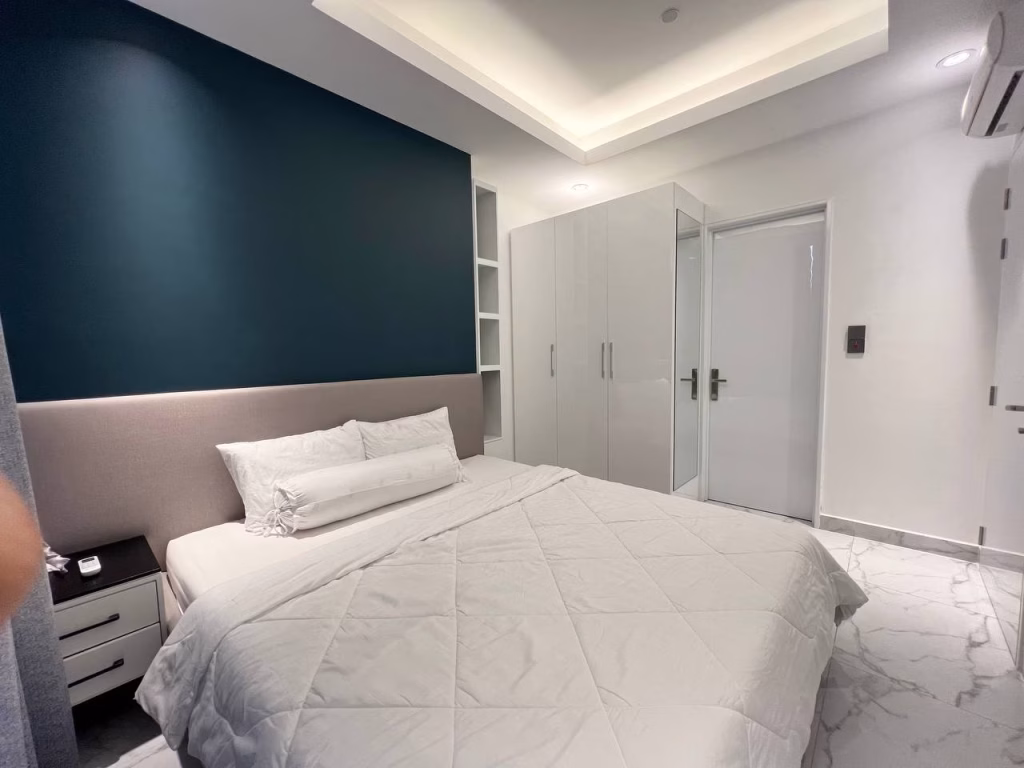 A modern 2 bedroom condo features a spacious bedroom with a large bed, white bedding, a dark blue accent wall, built-in wardrobes, marble floor, recessed ceiling lighting, and a nightstand with a small item on it.
