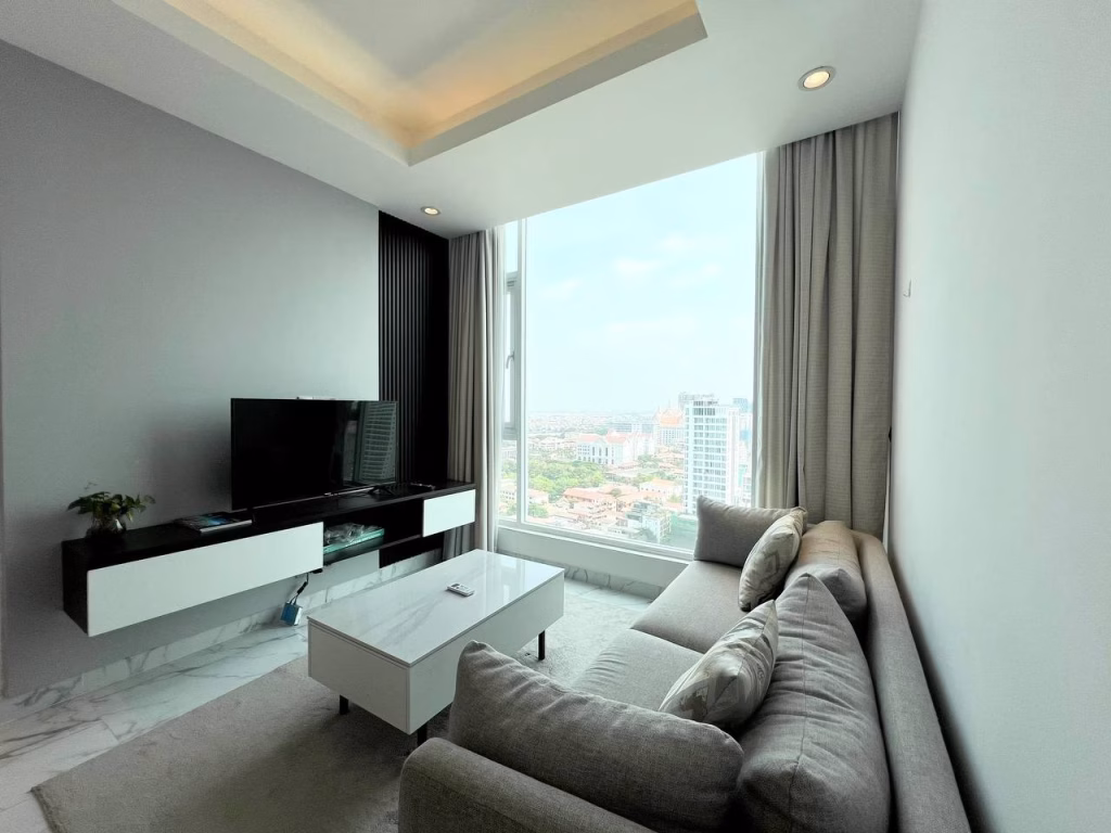 A modern living room in a 2 bedroom condo features a gray sofa, white coffee table, wall-mounted TV, and large window with city views. Light fills the space, highlighting minimalist decor and neutral tones.