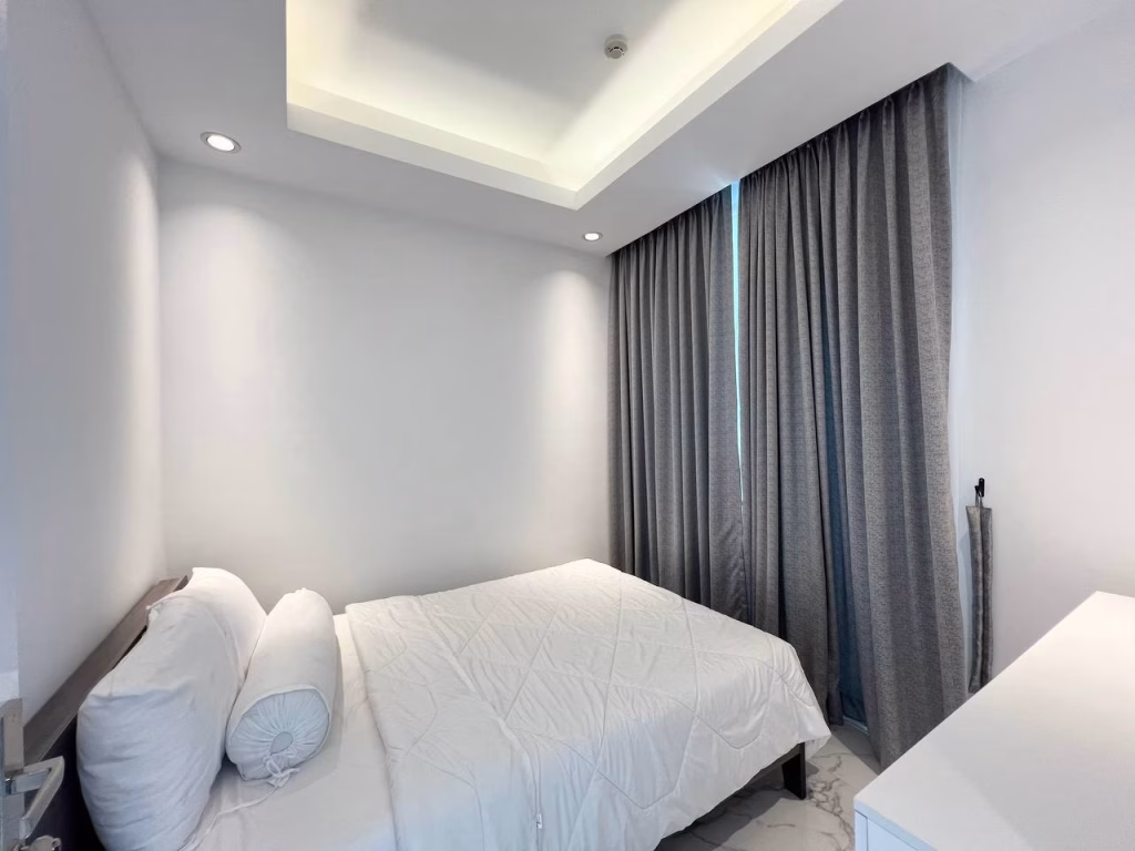 A modern, minimalistic 2 bedroom condo bedroom with a neatly made bed, white walls, gray floor-length curtains, recessed ceiling lighting, and a white dresser in the corner.