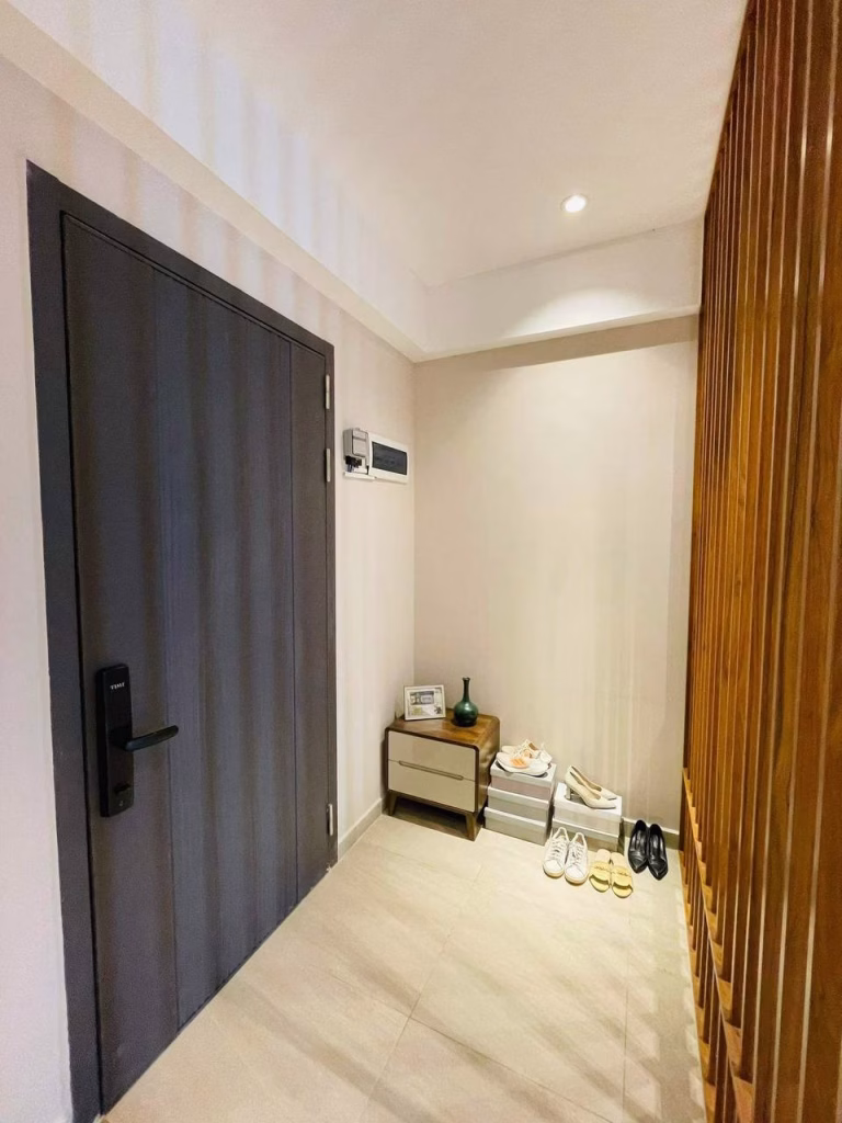 A modern apartment entryway features a dark door, a small wooden table with decor, neatly arranged shoes, and a partial wooden slat divider. The space is bright and minimalistic—perfect for those searching for an Apartment For Sale at this Time.