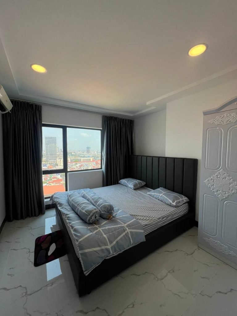A modern bedroom with a large bed, gray bedding, pillows, and rolled blankets. The room has marble floors, a white wardrobe, dark curtains, and a window with a city view under a bright sky.
