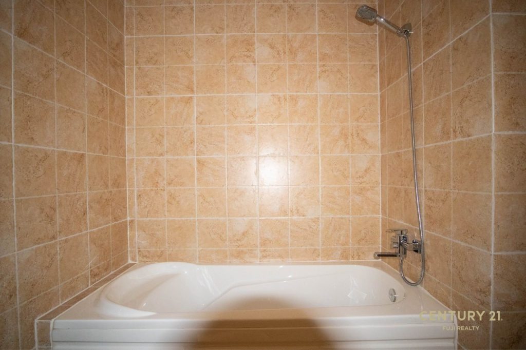 A white bathtub with a wall-mounted showerhead is set against tan, square tiled walls in a bathroom. The tiles have a warm, earthy tone, and the shower fixtures are chrome.