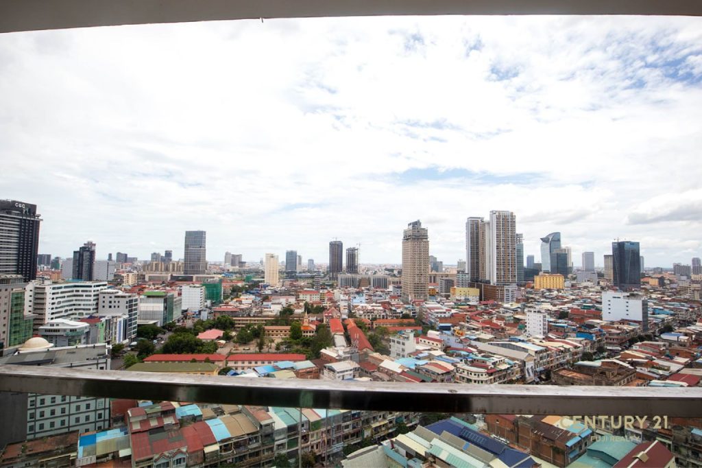A cityscape view from a high-rise balcony at Olympia city, Phnom Penh, showing densely packed buildings, skyscrapers, and cloudy sky above.