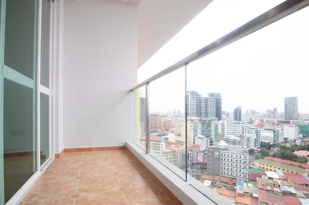 A modern high-rise balcony with brown tiled flooring, glass railing, and a view of a cityscape with tall buildings and cloudy sky.