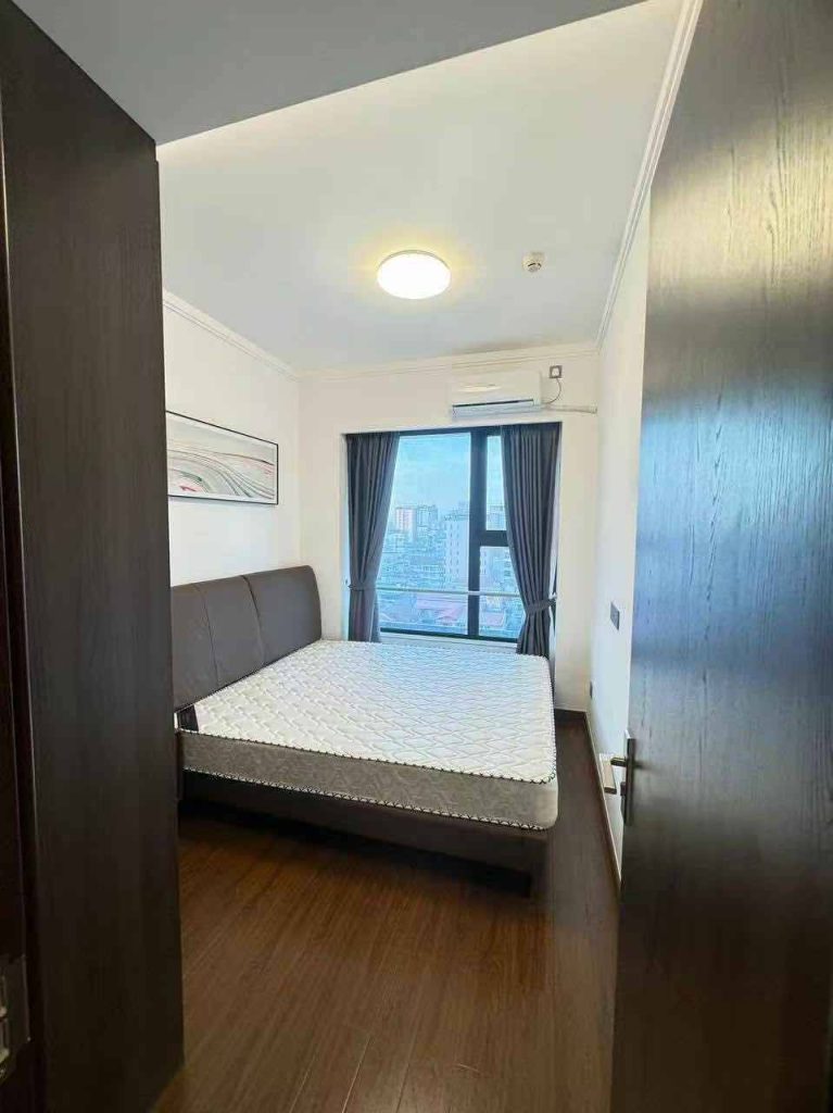 A small, modern bedroom with a double bed, dark headboard, wooden floor, large window with city view, blue curtains, air conditioner, and a round ceiling light. The door is partially open.