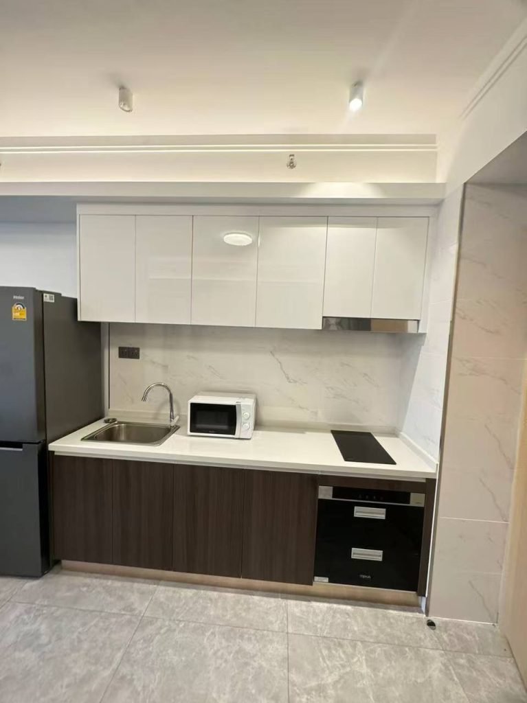 Modern kitchen with white upper cabinets, dark lower cabinets, a sink, microwave, stovetop, oven, and a refrigerator on the left. The backsplash and countertop are white, and the floor is tiled.