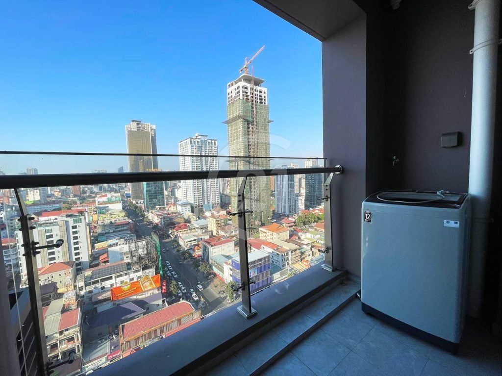 A city view from a high-rise balcony with glass railing, overlooking tall buildings and busy streets under a clear blue sky. A washing machine is placed in the corner of the balcony.