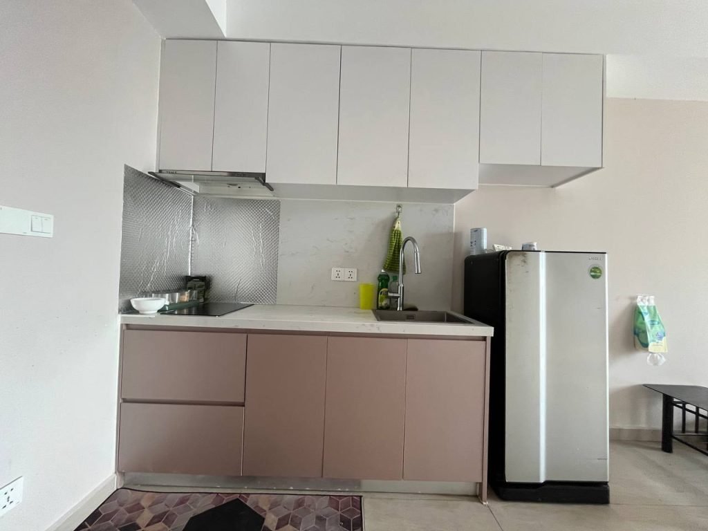 A modern kitchen with light brown lower cabinets, white upper cabinets, a stainless steel fridge, sink with a green scrubber, bowl on the counter, and a metallic backsplash behind the stove.