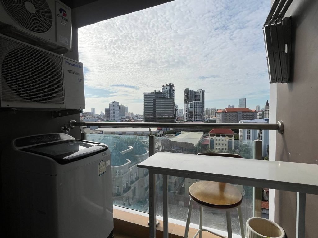 A small balcony with a washing machine, table, and stool overlooks a cityscape featuring tall buildings under a partly cloudy sky. An air conditioning unit is mounted on the wall above the washing machine.
