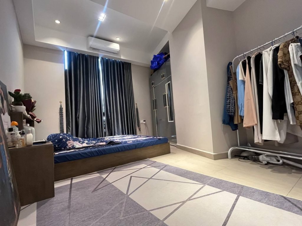 A modern bedroom with a low bed against the wall, dark curtains covering the window, an air conditioner above, a clothes rack with assorted clothing, and a patterned rug on the light-colored tiled floor.