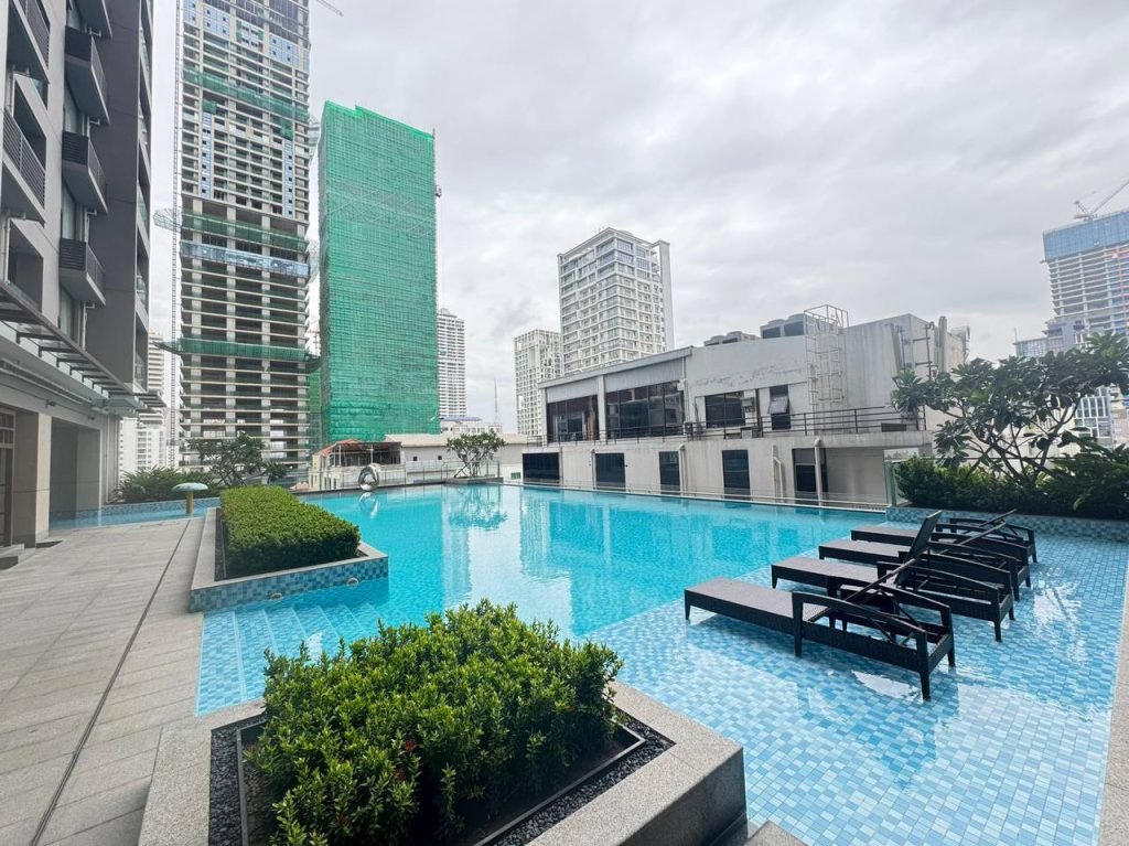 A modern rooftop swimming pool with lounge chairs, surrounded by city buildings and skyscrapers on a cloudy day. Greenery lines the poolside, and tall buildings are visible in the background.
