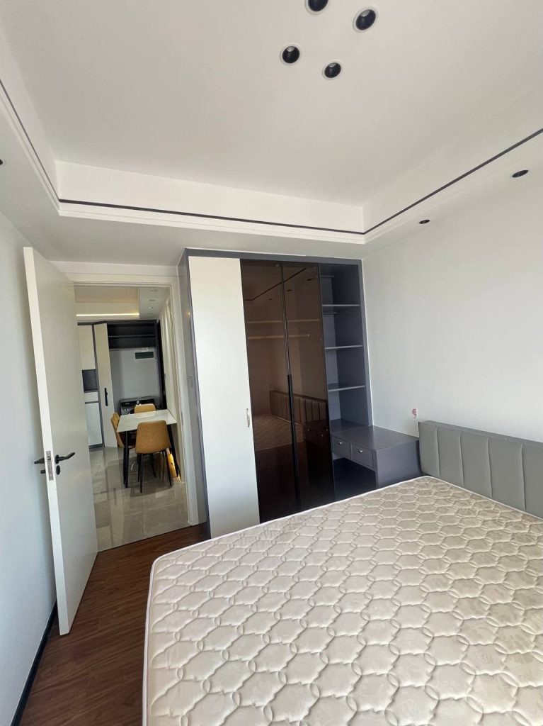A modern, bright bedroom with a bed, built-in wardrobe with glass doors, shelves, and a small desk. An open door reveals a dining area with a table and chairs in the background.