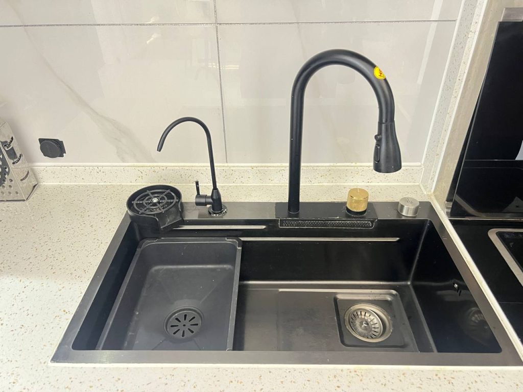 A modern kitchen sink with two basins, a black pull-down faucet, a smaller black filter tap, a soap dispenser, and a drain rack on a light-colored countertop with white tiled backsplash.