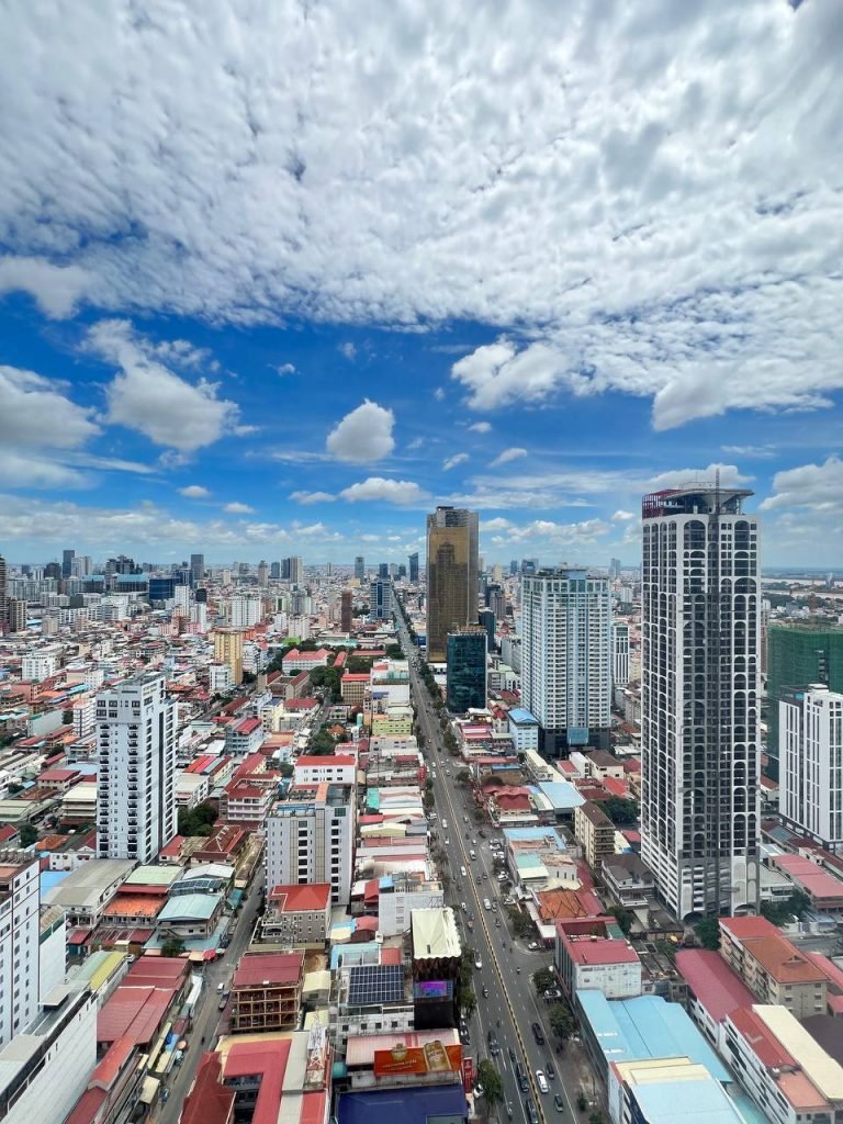 A vibrant cityscape featuring tall skyscrapers and busy streets under a bright blue sky with scattered clouds. The view captures urban buildings and a main road stretching toward the horizon.