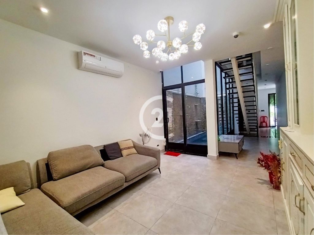 A modern living room with a gray sectional sofa, white walls, a large chandelier, and floor-to-ceiling windows near a staircase. The room has tiled floors and minimal decor.