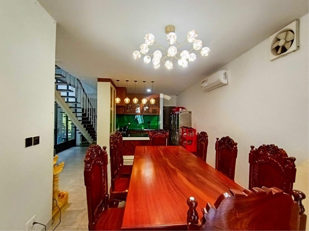 A modern dining room with a polished wooden table and ornate carved chairs. There’s a chandelier above, a staircase to the left, open kitchen with green backsplash, and white walls with air conditioner and exhaust fan.