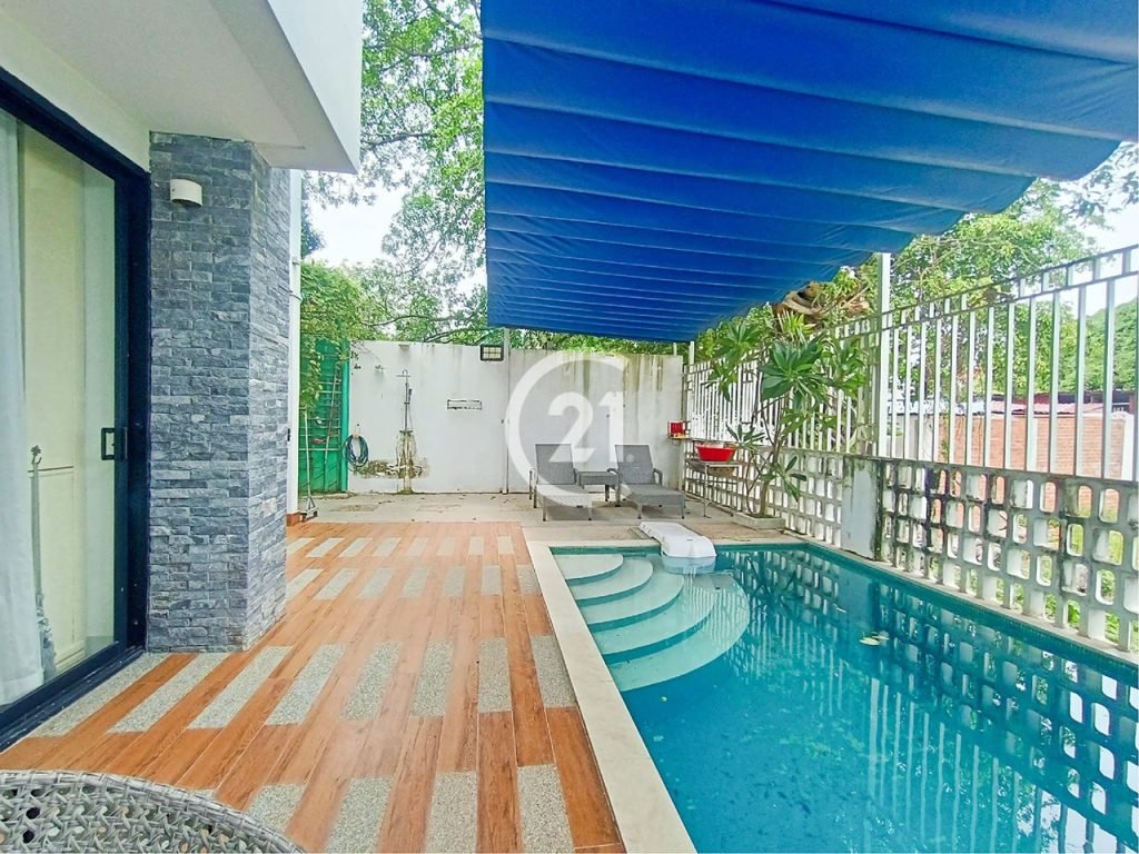 A small backyard pool with blue awning overhead, wooden and stone patio, pool steps, white lounge chairs, and a tall fence with greenery in the background.